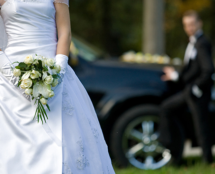 A bride holding a bouquet with a groom in the background near a black car.