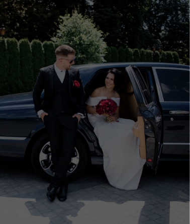 Bride and groom sitting by a black limousine, smiling.